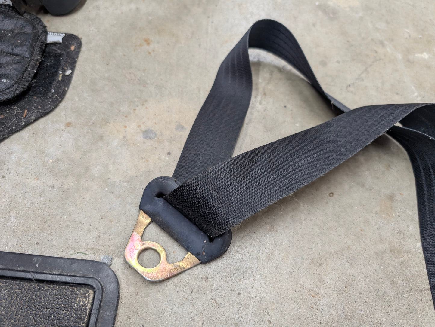 A General Motors NOS Passenger Seat Belt Retractor in charcoal for 1982-1993 Chevy S10 and Blazer rests on a gray concrete floor by the corner of a matching Chevrolet S10 floor mat.