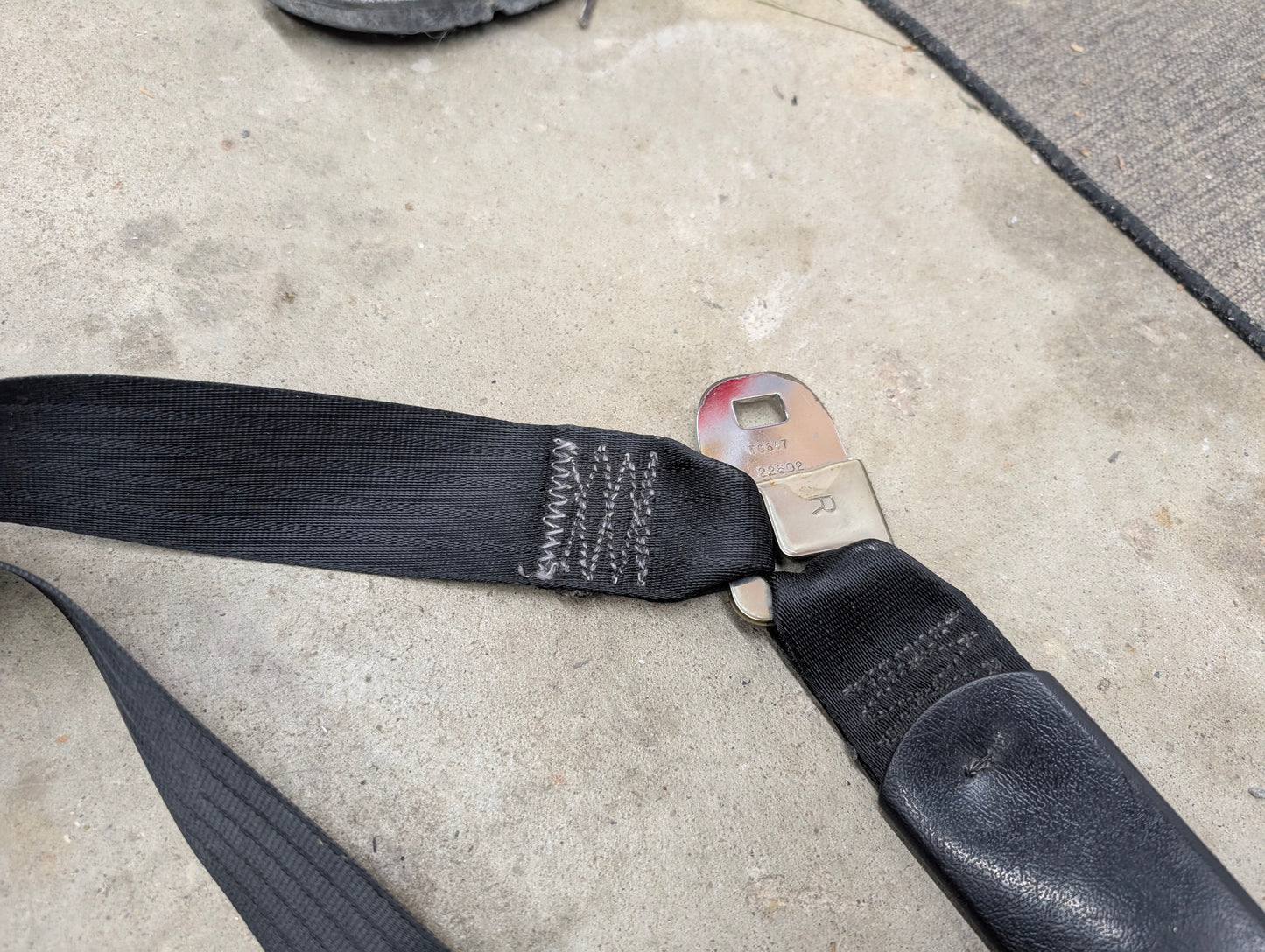 A close-up shows a General Motors NOS Passenger Seat Belt Retractor in Charcoal for 1982–1993 Chevy S10 and Blazer, partially extended with metal buckle, on concrete with floor mat and shoe visible in the background.
