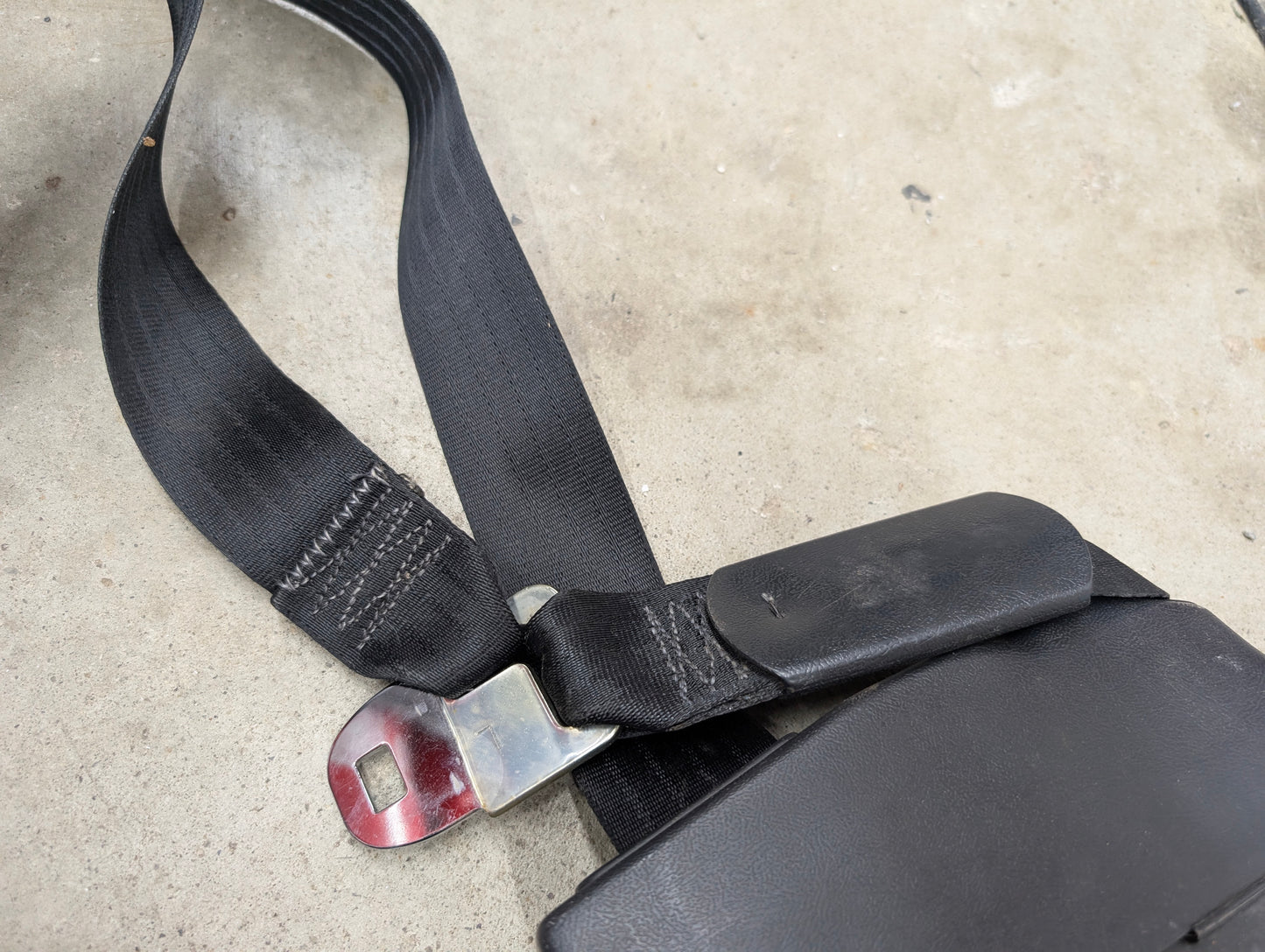 A close-up of a General Motors NOS Passenger Seat Belt Retractor in Charcoal for 1982–1993 Chevy S10, Blazer, and more, showing its black fabric texture, metal latch, and padded section on a concrete surface.