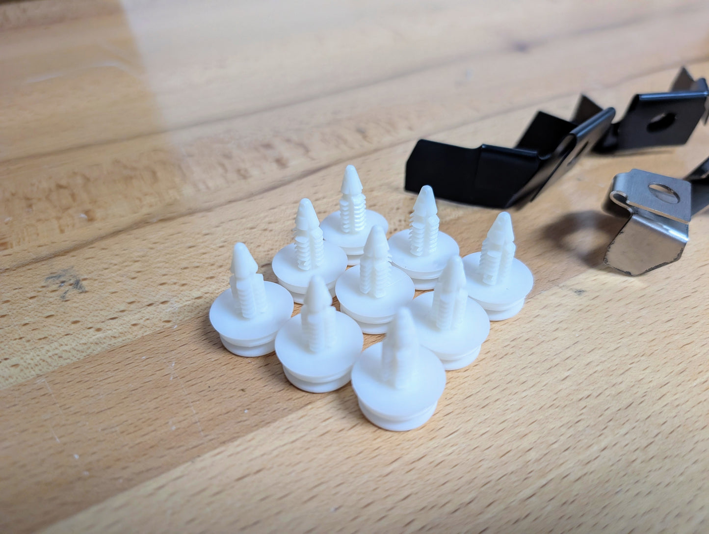 Nine white clips from The Ugly Truckling KIT Door Panel Armrest Repair for 1998-2005 Chevy S10, Blazer, GMC Sonoma & Jimmy are arranged on wood, with two black and one silver fasteners nearby.