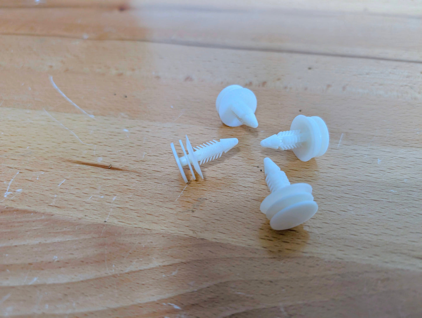 Four white plastic clips from The Ugly Truckling KIT Door Panel Armrest Repair w/Clips for 1998-2005 Chevy S10, Blazer, GMC Sonoma, and Jimmy rest on a lightly scratched wooden surface.