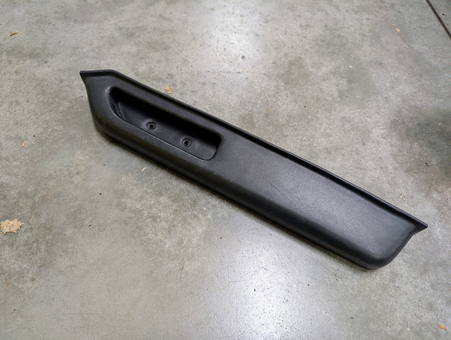 A General Motors OEM black passenger door panel armrest handle for 1986-1993 Chevy S10 Blazer, GMC Jimmy, and more rests on a gray concrete floor. The handle is rectangular with angular edges, recessed grip area, and two screw holes.