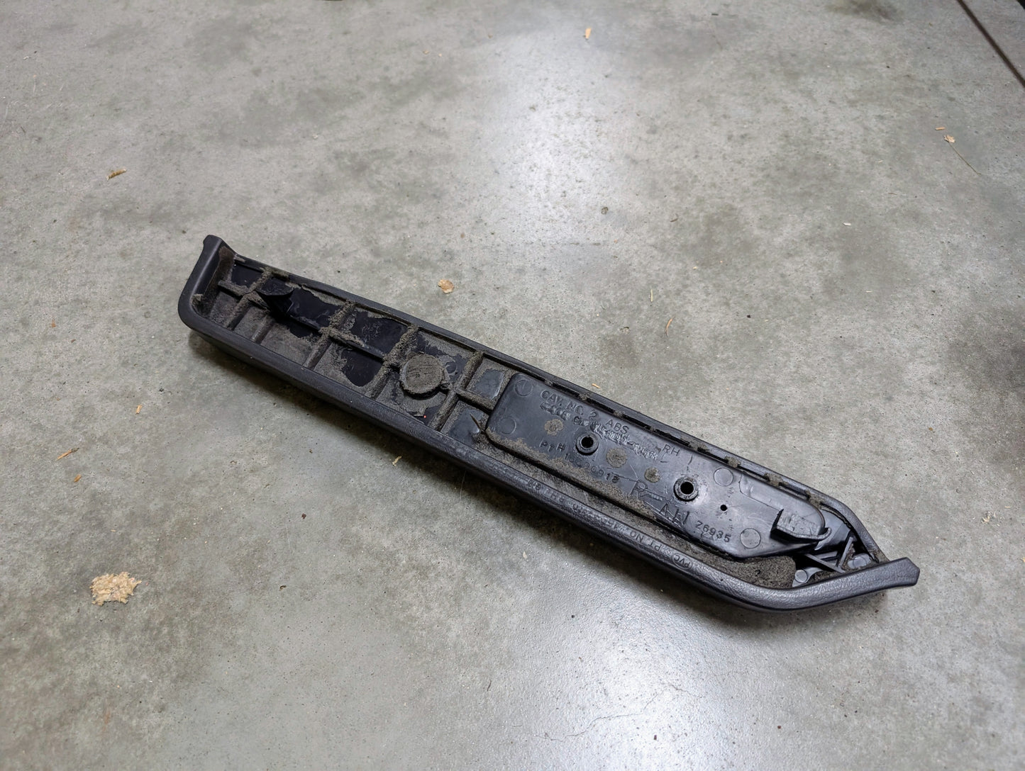 A General Motors OEM black passenger door panel armrest handle for 1986-1993 Chevy S10, Blazer, GMC Jimmy, and more rests on a gray concrete floor. The elongated part has an angled shape with a slightly curved end.