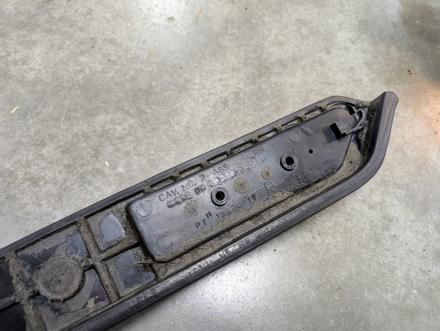 A close-up of an OEM black passenger door panel armrest handle by General Motors for 1986-1993 Chevy S10 Blazer and GMC Jimmy, resting on concrete and showing molded part numbers and manufacturing details.