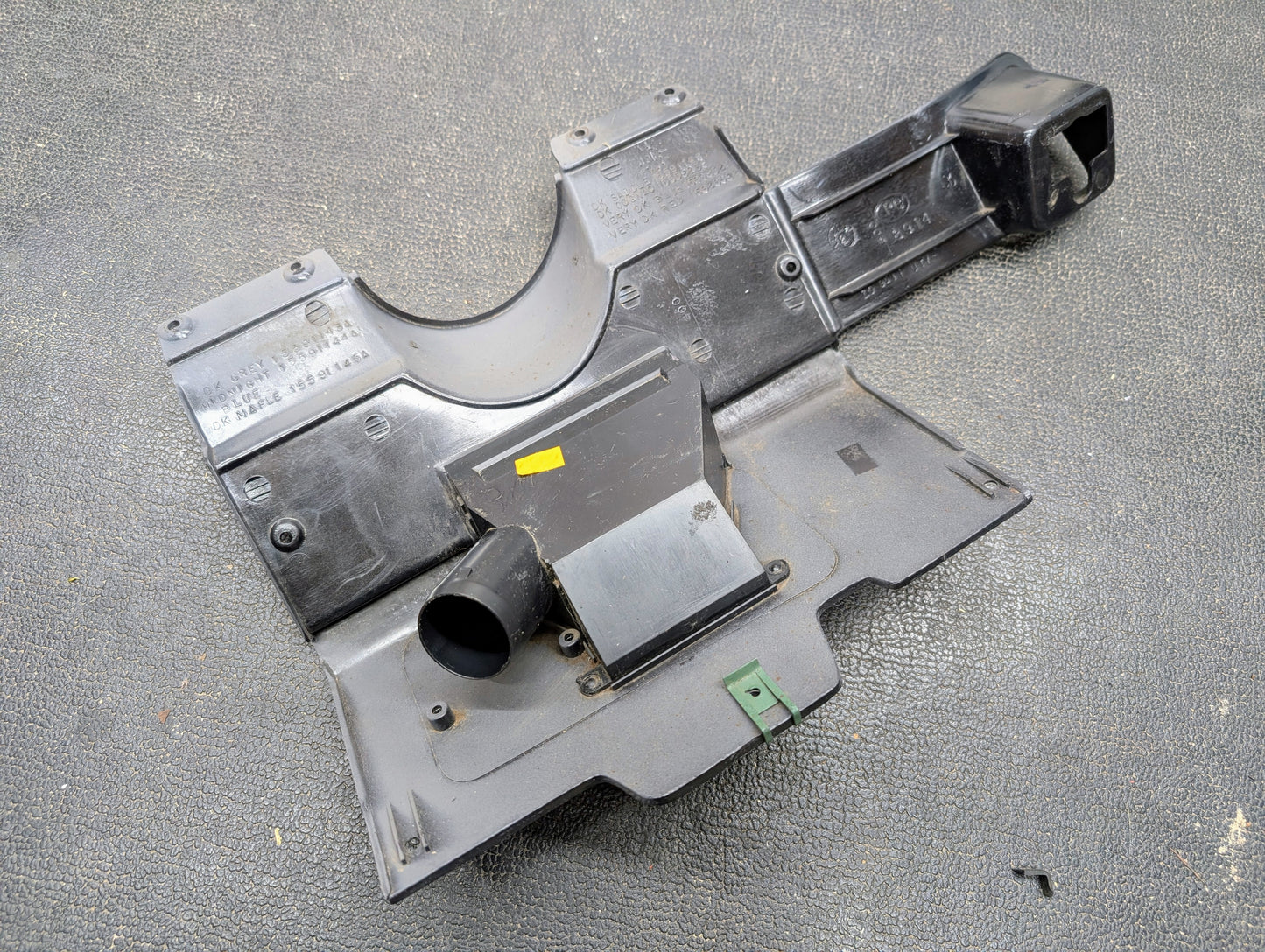 An OEM Lower Dash Trim Knee Bolster in Lt Gray for 1986-1993 Chevy S10 Blazer, GMC S15, and more by General Motors rests on a textured gray surface, featuring molded sections, mounts, a yellow sticker, and a green plastic piece.