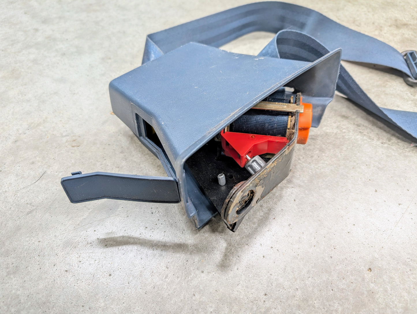 A close-up of an OEM General Motors blue driver seat belt retractor & buckle for 1982-1993 Chevy S10/GMC S15 with a broken plastic cover, exposing internal parts, lying on a light-colored concrete surface.
