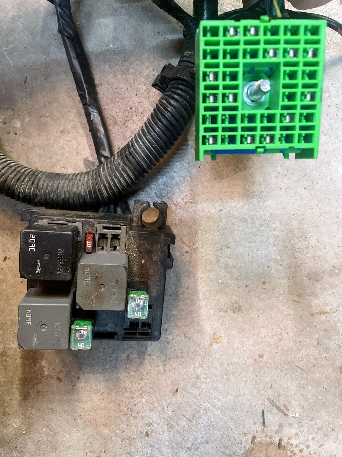 A close-up of the General Motors KIT Electric Fan Kit with Factory Harness for 1999-2006 GM Full Size Trucks, showing multiple relays, fuses, and a green connector—perfect for fan upgrades—resting on concrete.