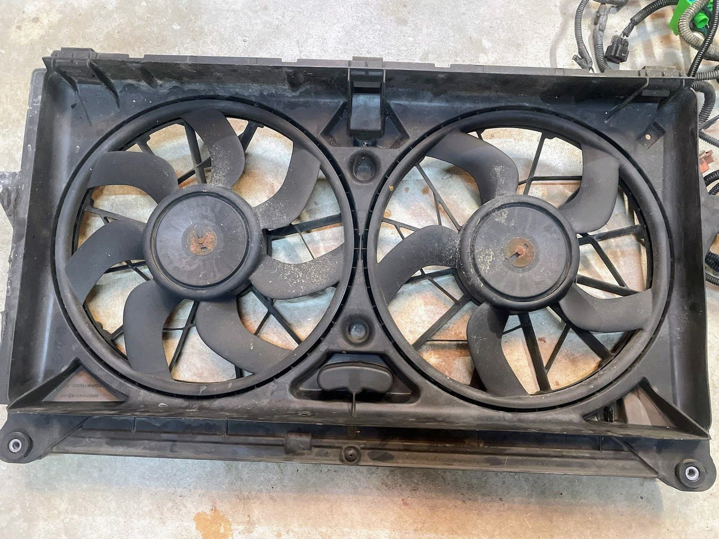 A close-up of the General Motors KIT Electric Fan Kit with Factory Harness for 1999-2006 GM Full Size Trucks, showing two large black fans in a rectangular shroud with some dust and dirt on the surfaces.