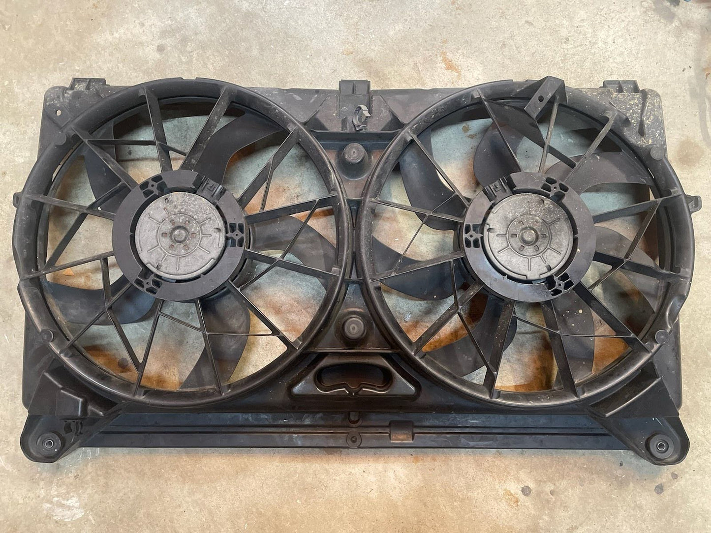 A General Motors KIT Electric Fan Kit with Factory Harness for 1999-2006 GM Full Size Trucks, featuring large black fans on a rectangular frame, is shown resting on concrete with visible rust and wear in the metal background.