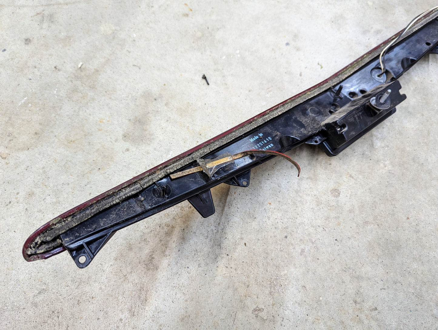 A close-up of a General Motors OEM Center High Mount Third Brake Lamp (CHMSL) for 2003-2007 Cadillac CTS shows exposed wires and internal components on a concrete surface, with the assembly detached to reveal mounting brackets.