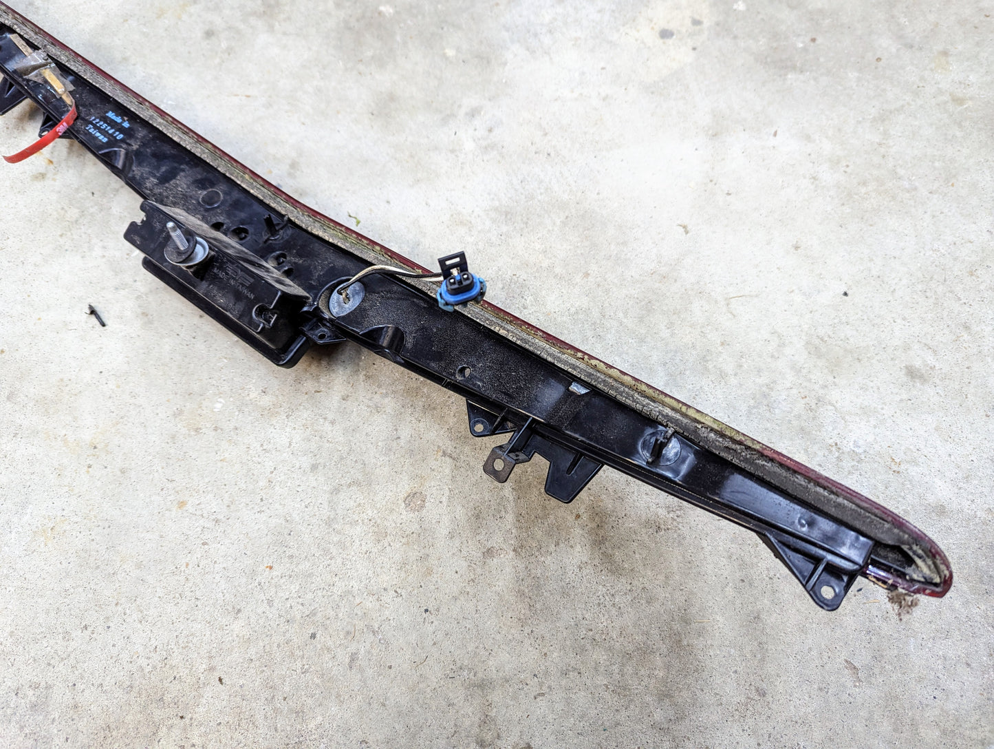 A General Motors OEM Center High Mount Third Brake Lamp (CHMSL) for 2003-2007 Cadillac CTS, in black plastic with wiring, screws, clips, and a blue electrical connector, is shown lying on a concrete floor.