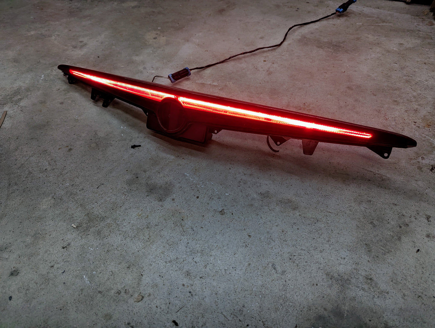 A General Motors OEM Center High Mount Third Brake Lamp (CHMSL) for 2003-2007 Cadillac CTS, illuminated in red with an integrated rearview camera, rests on a concrete floor with its power cable trailing behind.