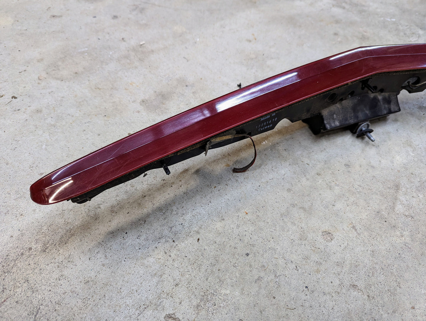 A General Motors OEM Center High Mount Third Brake Lamp (CHMSL) for 2003-2007 Cadillac CTS, with visible wiring and a slightly scratched red lens, rests detached on a light gray concrete surface.