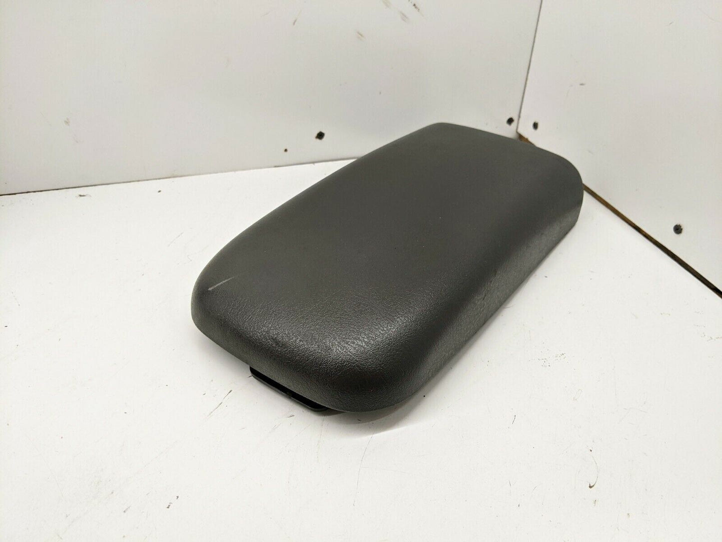 A General Motors 02-09 GMC Envoy Chevy Trailblazer dark gray center console armrest lid is shown on a white surface against a white wall, displaying minor scuff marks and blemishes.