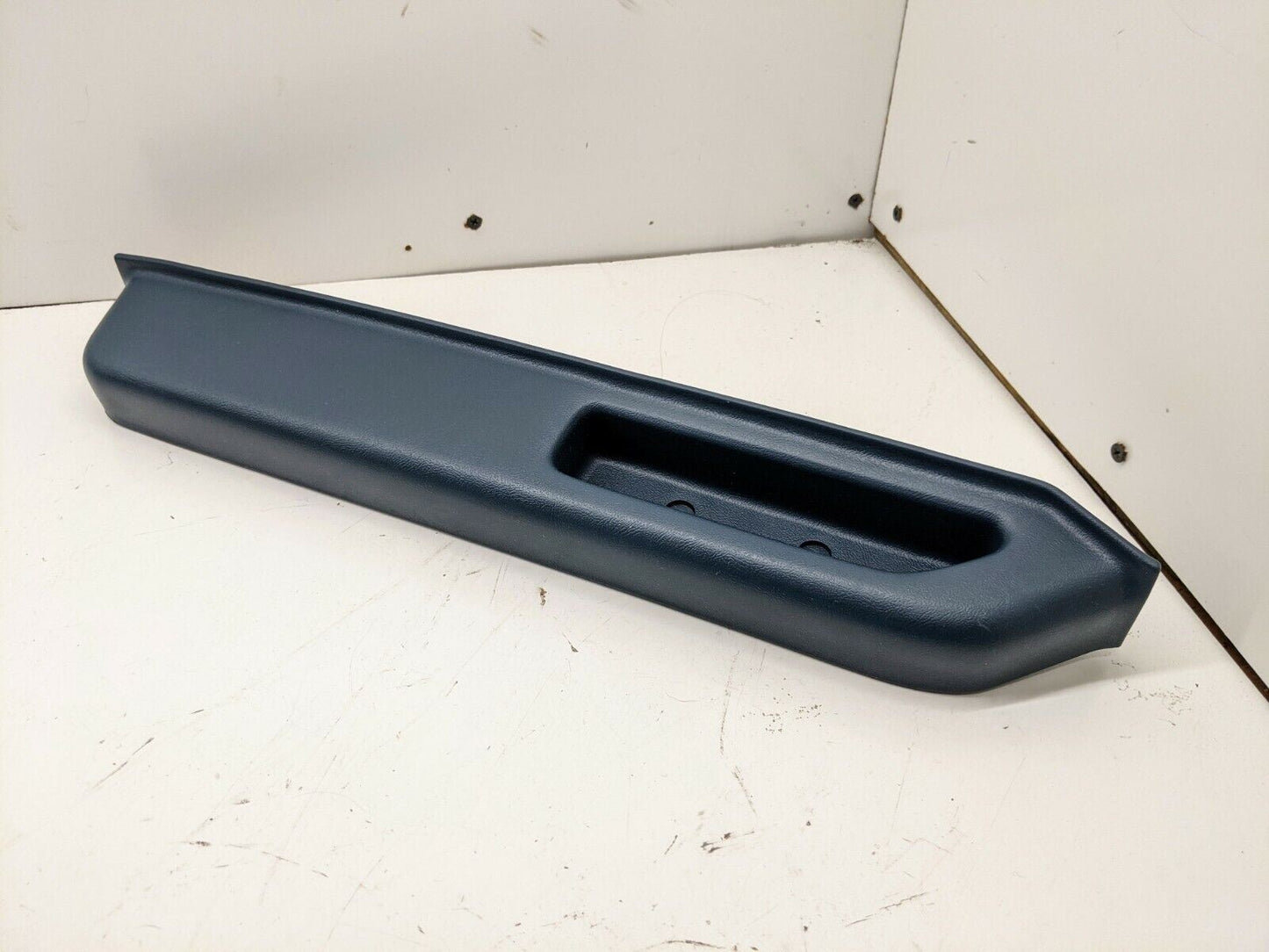 A General Motors blue plastic NOS door panel armrest handle for 1986-1993 Chevy S10 Blazer or GMC Jimmy, shown on a white surface against two white walls.