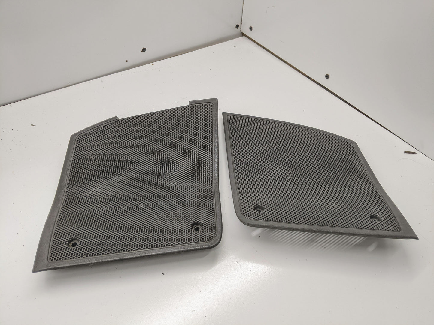 A pair of General Motors OEM light gray dash speaker grille covers for 1995-1997 Chevrolet S10 and Blazer are shown on a white surface, leaning against a white corner wall.