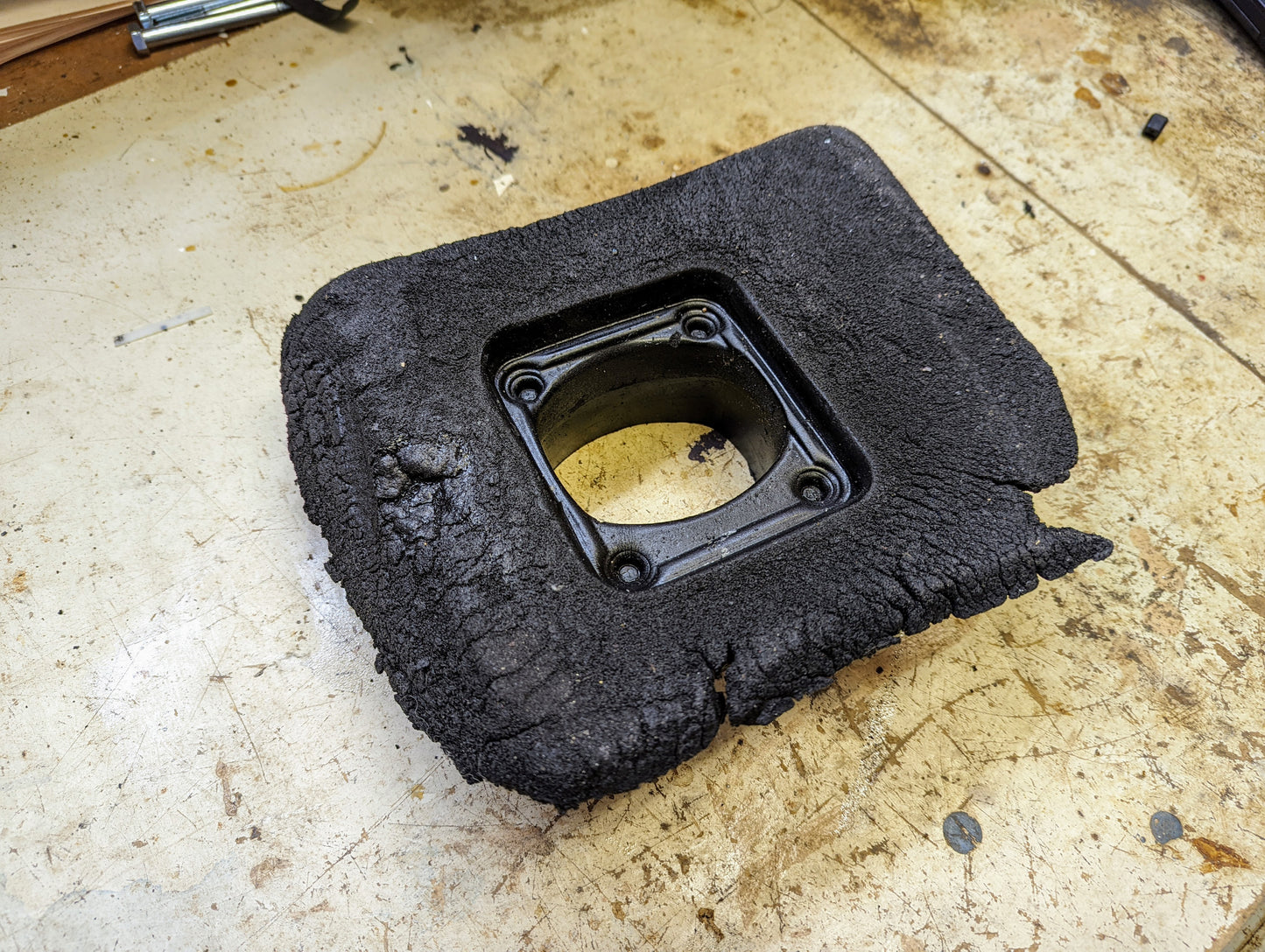 A heavily charred, blackened Manual Floor Shifter Insulator Foam for 1998-2005 Chevy S10 Blazer GMC Sonoma Jimmy by General Motors rests on a stained, worn workbench, featuring a metal insert and four screws.