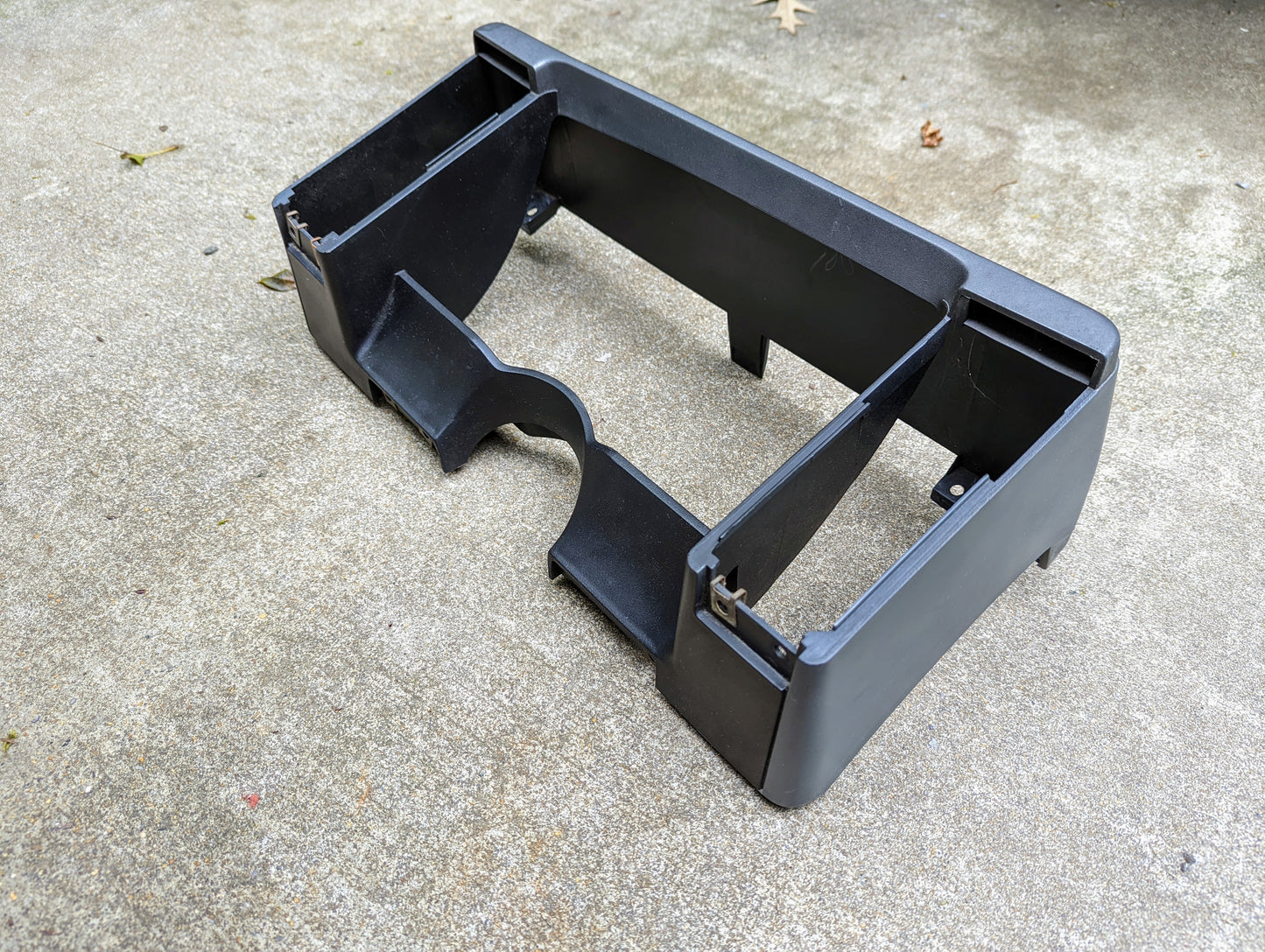A gray General Motors OEM Instrument Cluster Dash Bezel for 1986–1993 Chevy S10, Blazer, and GMC S15 is shown on rough concrete. It features curved and straight compartments plus metal clips on the sides.