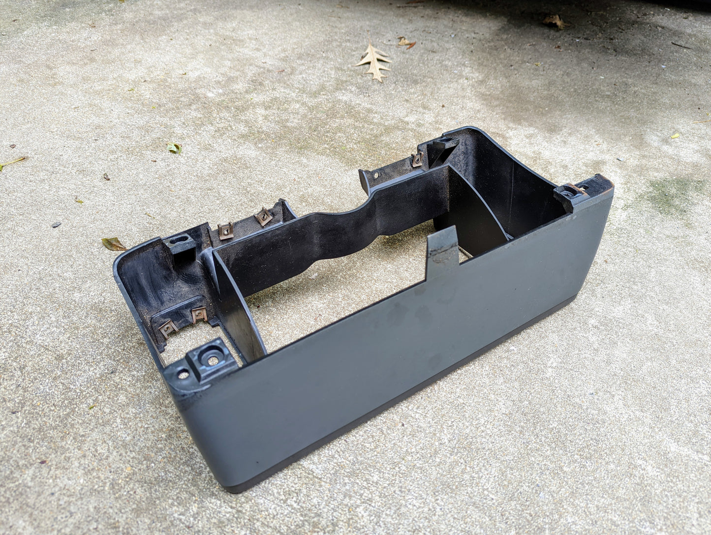 A gray General Motors OEM instrument cluster dash bezel for 1986-1993 Chevy S10, Blazer, GMC S15, and more rests on concrete. Some metal clips are visible inside, with a dry leaf nearby on the ground.