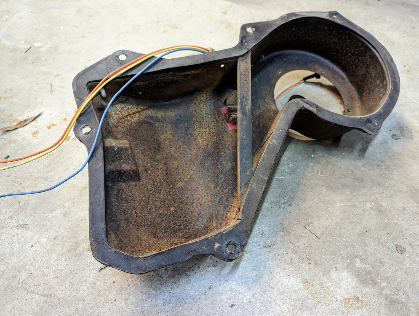 A rusty OEM Heater Box Non AC Blower Housing (General Motors) for 1982-1993 Chevy S10/Blazer, featuring rounded edges, mounting holes, and colored wires, rests empty on a concrete floor.