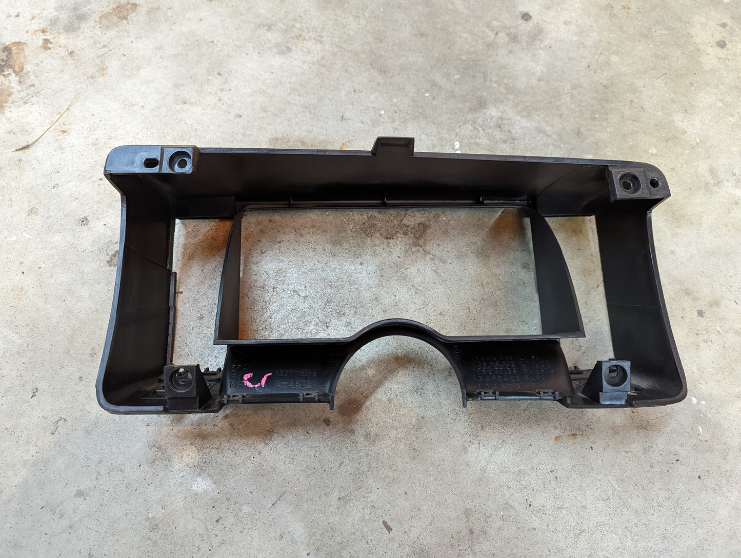 A General Motors OEM Instrument Cluster Dash Bezel in tan for 1986-1993 Chevy S10, Blazer, GMC S15, and more is shown resting on a concrete floor. This trim piece features an arched lower edge and multiple mounting points.