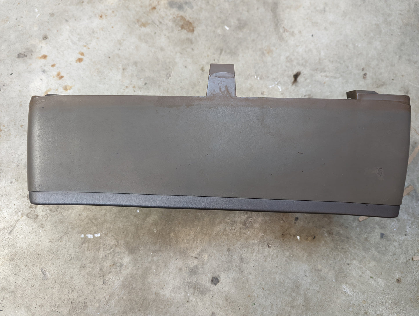 A tan OEM Instrument Cluster Dash Bezel by General Motors for 1986-1993 Chevy S10, Blazer, and GMC S15 rests on a rough, stained concrete surface, featuring a small raised tab on top.