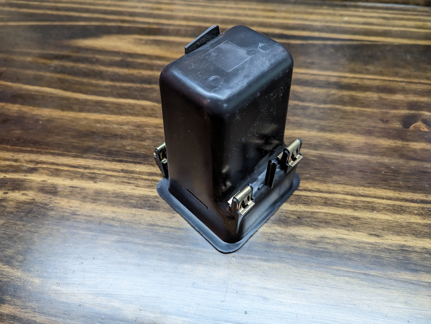 A black rectangular relay with metal prongs rests on a dark, striped wooden surface—similar to parts in the General Motors OEM Dash Pocket with Rubber Bin for 1999-2006 GM Full Size Trucks.