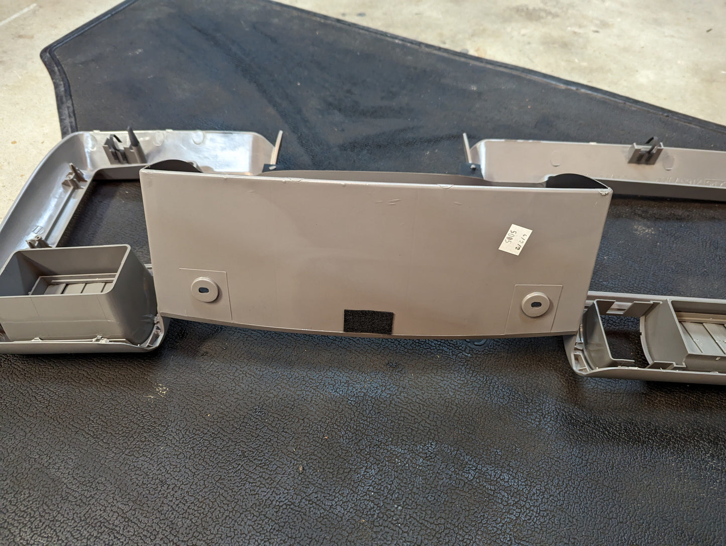 A NOS gray plastic General Motors instrument cluster dash bezel for 1995-1997 Chevy S-10, Blazer, and more is shown on a textured black surface with cup holder sections, mounting points, and a small white sticker visible.