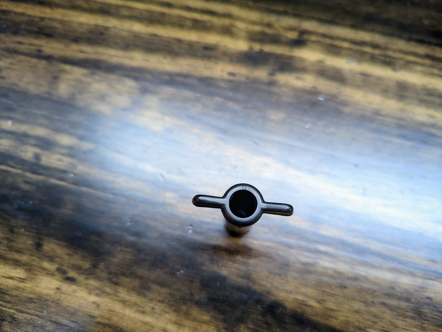 A black OEM wing nut by General Motors for 4.3/5.0/5.7L 1996-2007 GM Trucks rests on a striped wooden surface; its cylindrical, symmetrical shape with two horizontal extensions is seen from above.