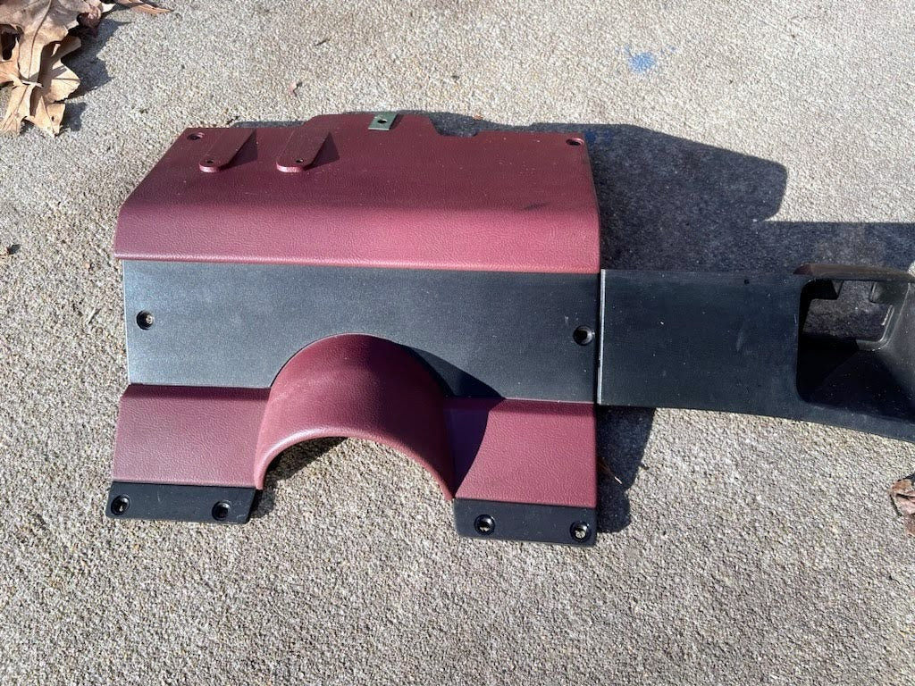 A General Motors OEM red lower dash trim knee bolster for 1986–1993 Chevy S10 Blazer, GMC S15, and more rests on concrete with dried leaves nearby. The curved plastic part has screw holes and protects the vehicle cabin.