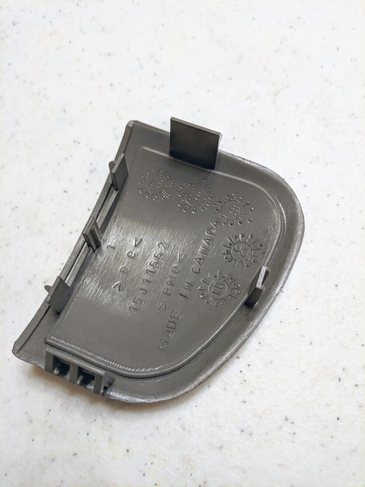 A close-up of a General Motors OEM 4x4 Delete Filler Plate Dash Blank Panel in gray for 1998-2005 Chevy S10, Blazer, and more, embossed with symbols and numbers, resting on a white speckled surface.