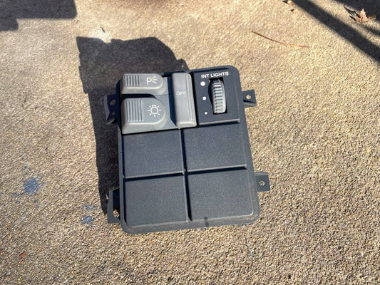 A General Motors OEM Headlight Headlamp Switch panel for 1994–1997 Chevy S10, Blazer, GMC Sonoma, and Jimmy rests on rough concrete in sunlight, with blank slots at the bottom and switches for parking lights, headlights, and interior lights.