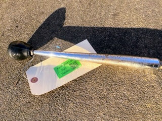 A General Motors OEM 5 Speed Manual Transmission Shifter Handle for 1982–1993 Chevy S10 and Blazer with a black round end rests on pavement beside a large white tag with a green label; both cast shadows in sunlight.