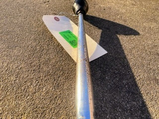 A close-up of a General Motors OEM 5 Speed Manual Transmission Shifter Handle for 1982-1993 Chevy S10, Blazer, and more, with a black knob, shows it lying on concrete. A white tag with green sticker is attached near the rod’s end. Shadows are visible.