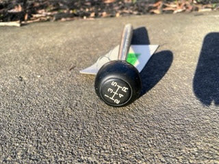 A close-up photo of a General Motors OEM 5 Speed Manual Transmission Shifter Handle for 1982-1993 Chevy S10 and Blazer, showing the shift pattern on its black knob, rests on a concrete surface in sunlight.