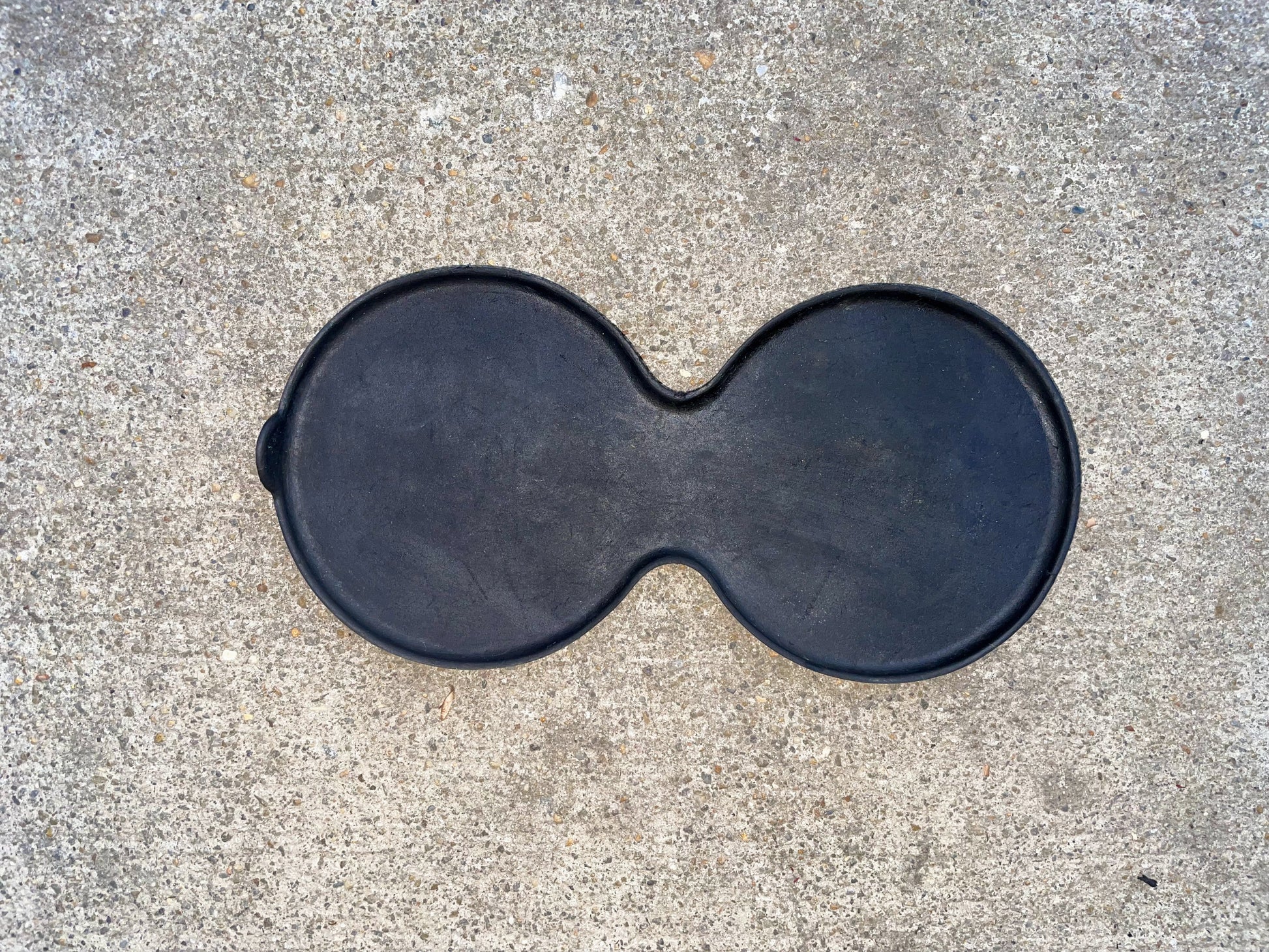 A black, figure-eight-shaped griddle rests on rough light gray concrete, resembling the General Motors OEM Center Console Cup Holder Bin Rubber Mat Liner for 2003-2006 Chevy Silverado and GMC Sierra.