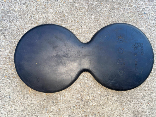 A black, figure-eight-shaped General Motors OEM Center Console Cup Holder Bin Rubber Mat Liner for 2003-2006 Chevy Silverado and GMC Sierra rests on rough concrete, showing indented text and small sun/snowflake symbols.