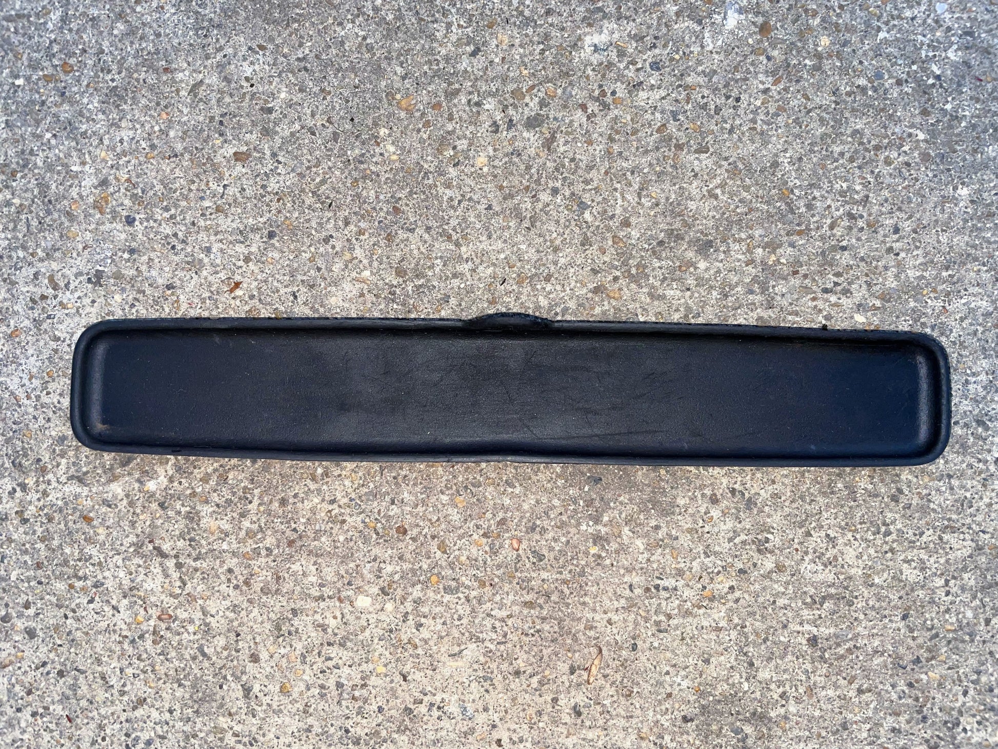 A black, rectangular rubber mat with rounded corners lies on speckled concrete; it resembles the General Motors OEM Center Console Cup Holder Central Storage Bin Rubber Mat Liner for 2003-2006 Chevy Silverado, GMC Sierra, and similar models.
