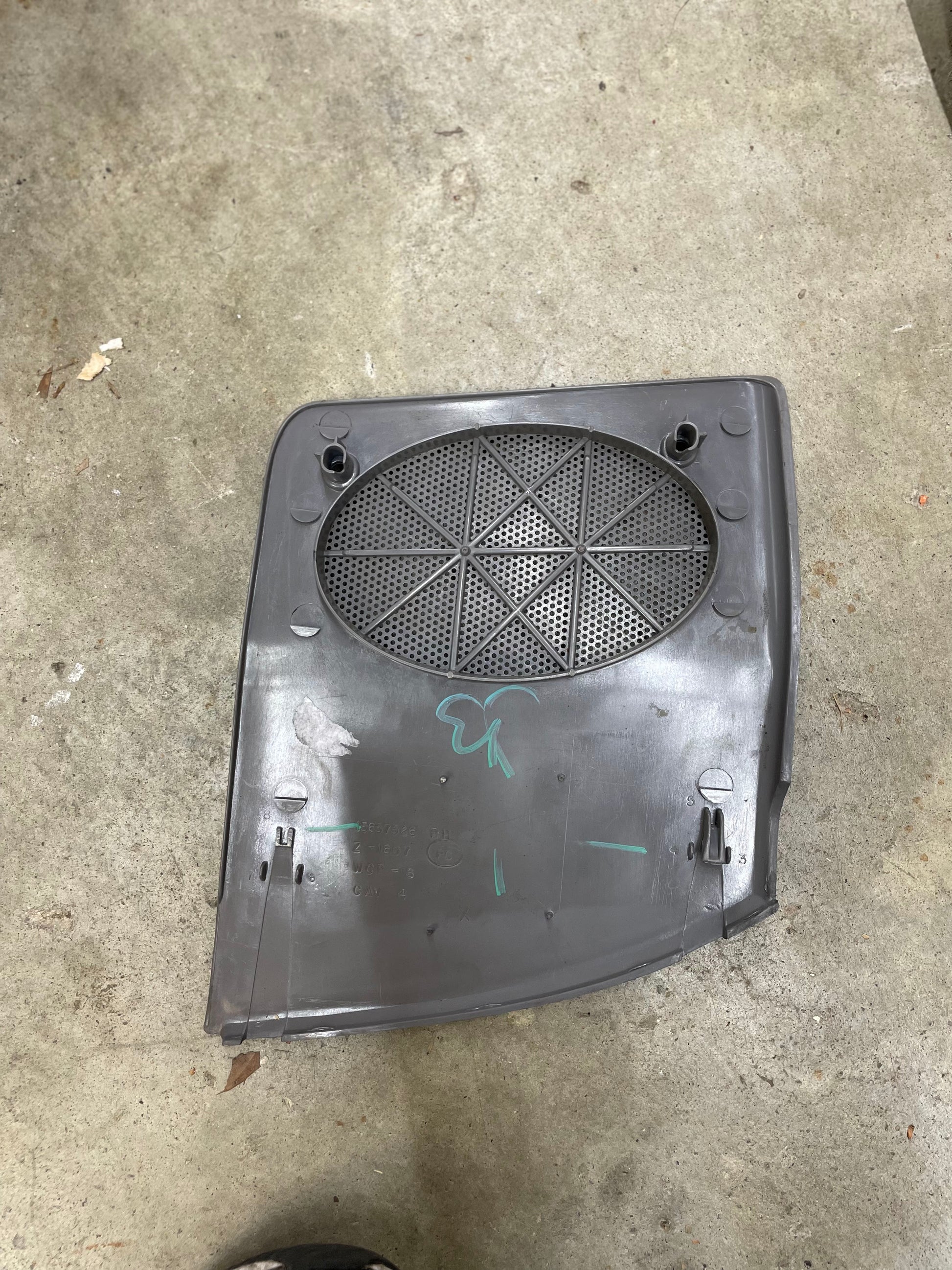 A General Motors OEM passenger RH dash speaker grille cover in light gray with a broken tab for 1995-1997 Chevrolet S10, Blazer, and more rests on concrete. Green markings and a cloverleaf are visible on the panel’s surface.