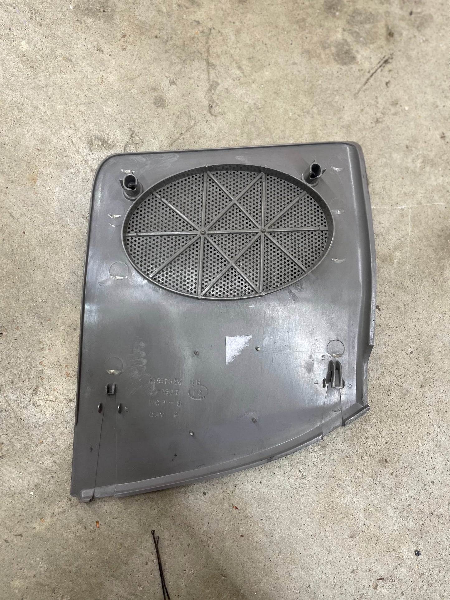 A General Motors OEM passenger RH dash speaker grille cover in light gray with a broken tab for 1995-1997 Chevrolet S10, Blazer, and more sits on concrete. This plastic panel has a circular perforated grille and multiple attachment points.