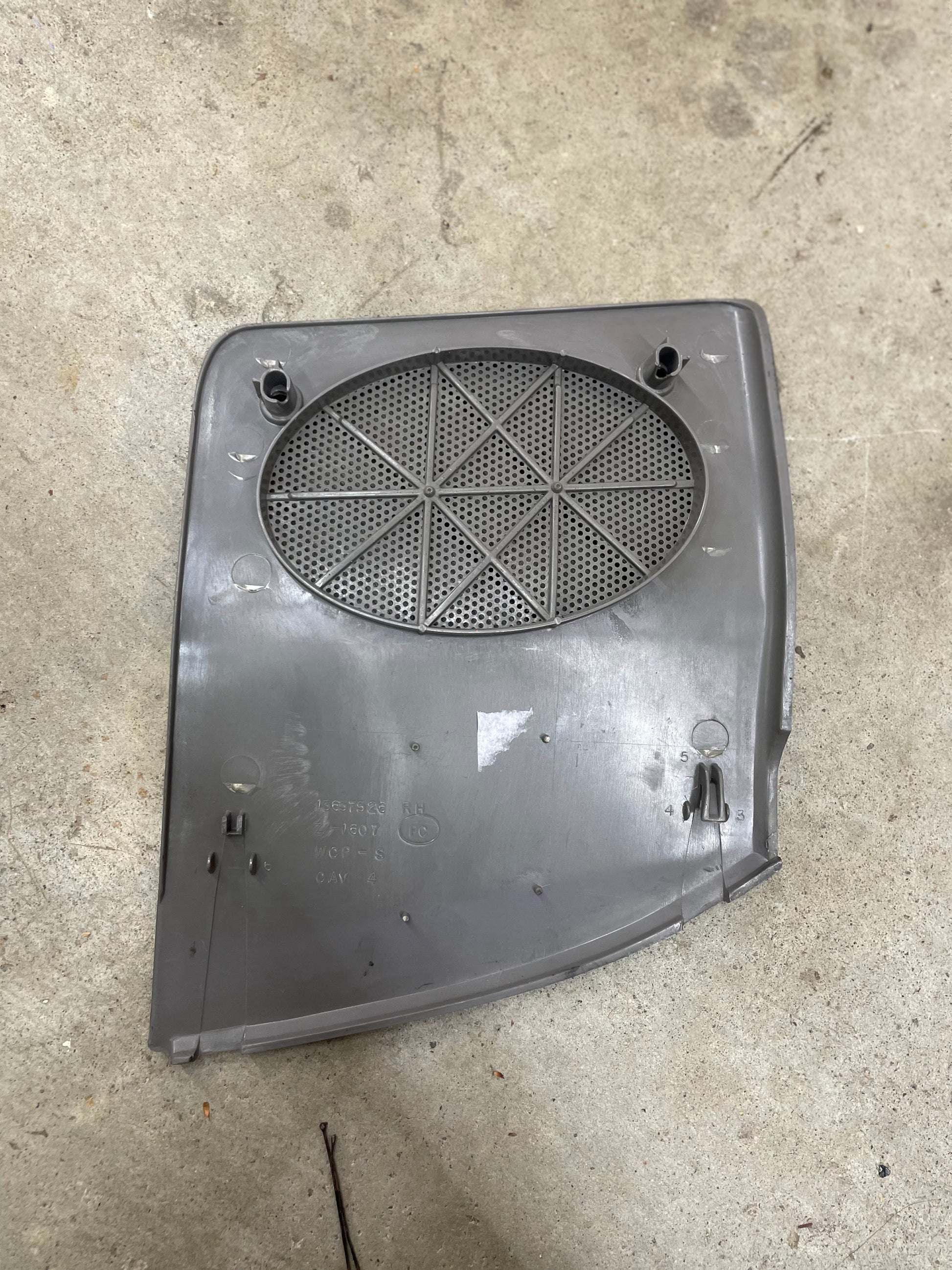 A General Motors OEM passenger RH dash speaker grille cover in light gray with a broken tab for 1995-1997 Chevrolet S10, Blazer, and more sits on concrete. This plastic panel has a circular perforated grille and multiple attachment points.