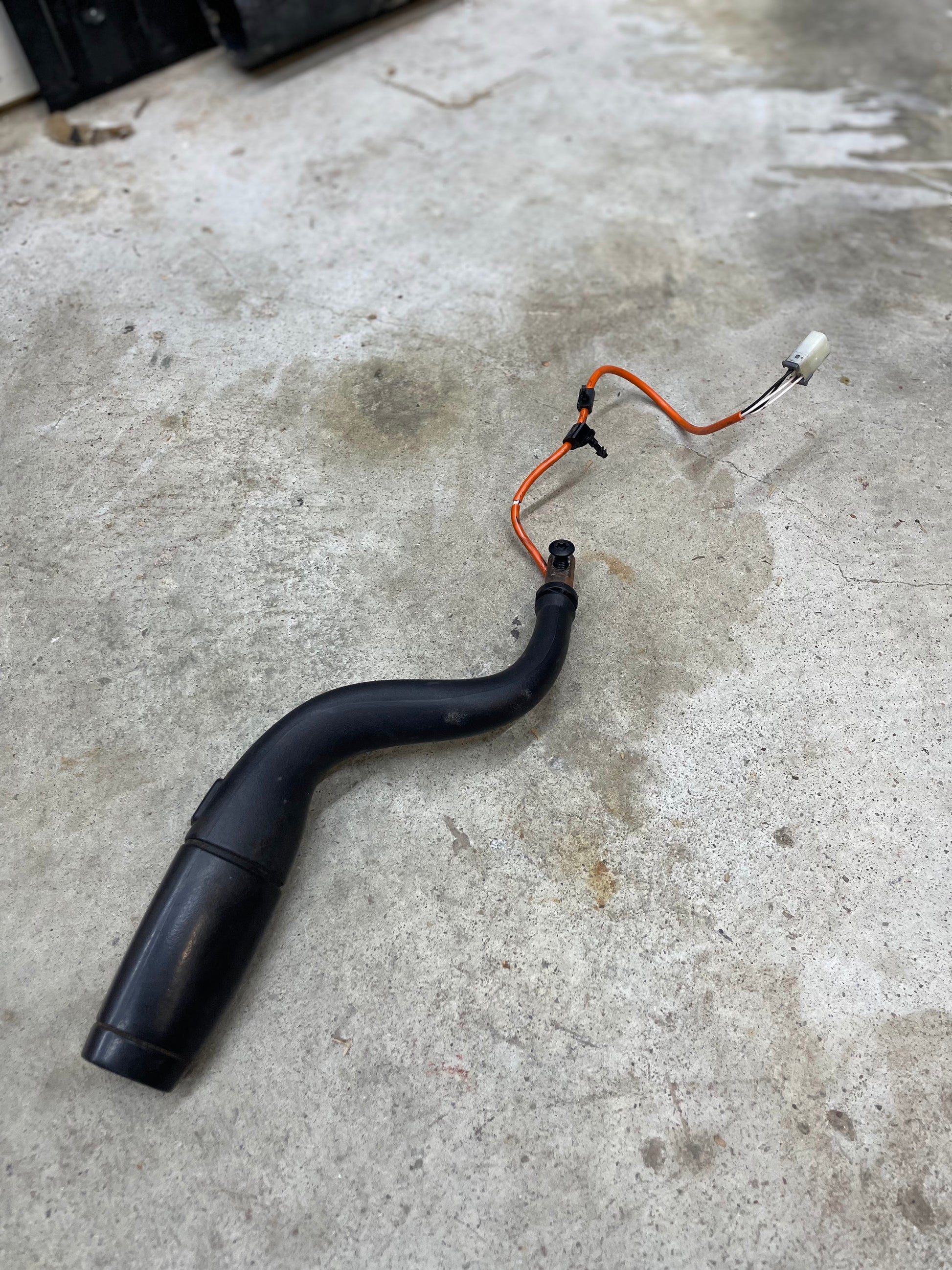 An OEM Column Shift Lever with Tow/Haul & Tapshift for 2006-2007 GM Duramax LBZ by General Motors, featuring a black curved pipe, orange and black wires, and a white connector, lies on a stained concrete floor.