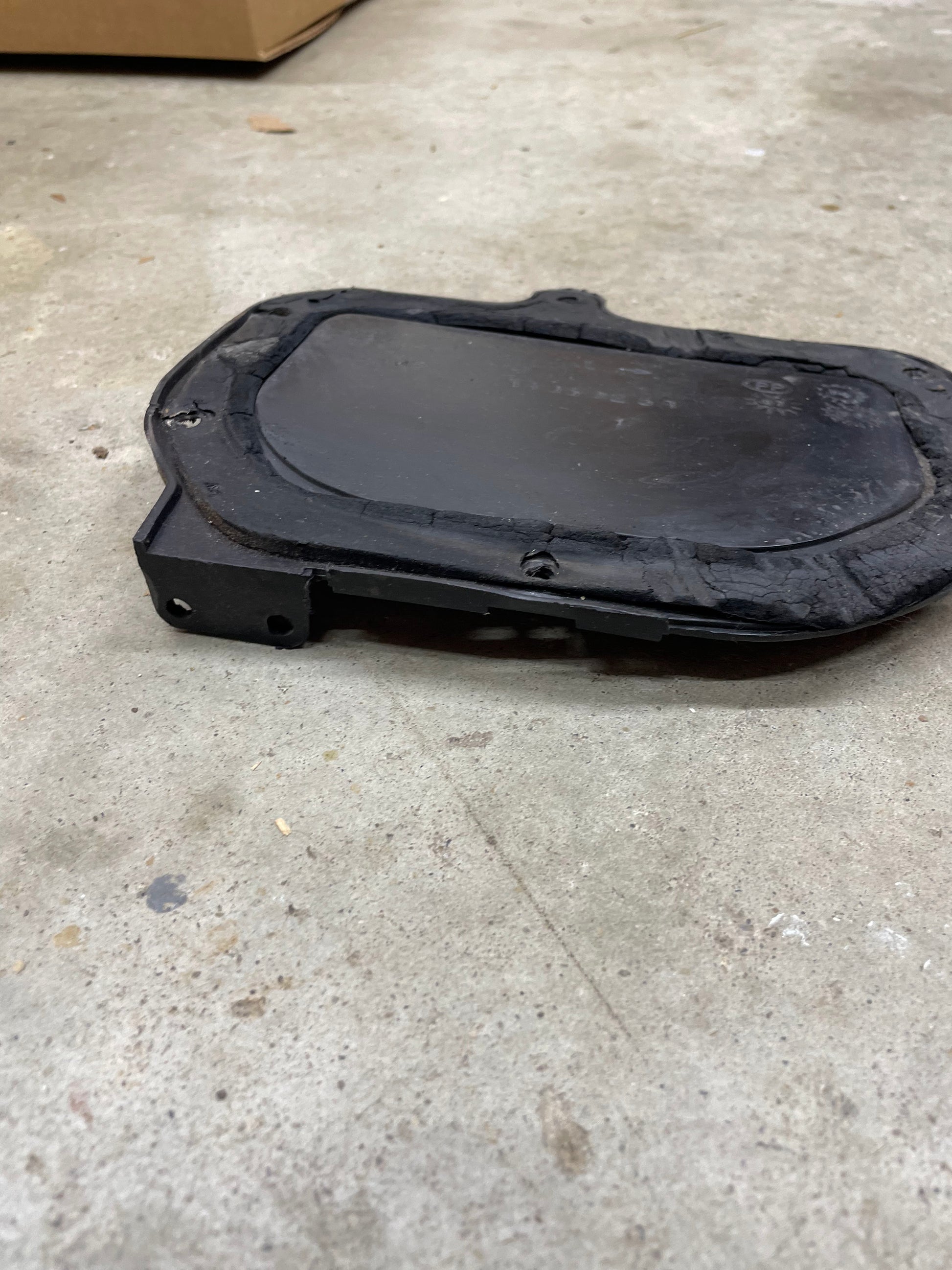 A black OEM Fresh Air Block-Off Panel by General Motors for 1982–1993 Chevrolet S10, Blazer, and more lies on a concrete floor. Slightly worn with screws and a raised border, it hints at prior use in automotive settings.