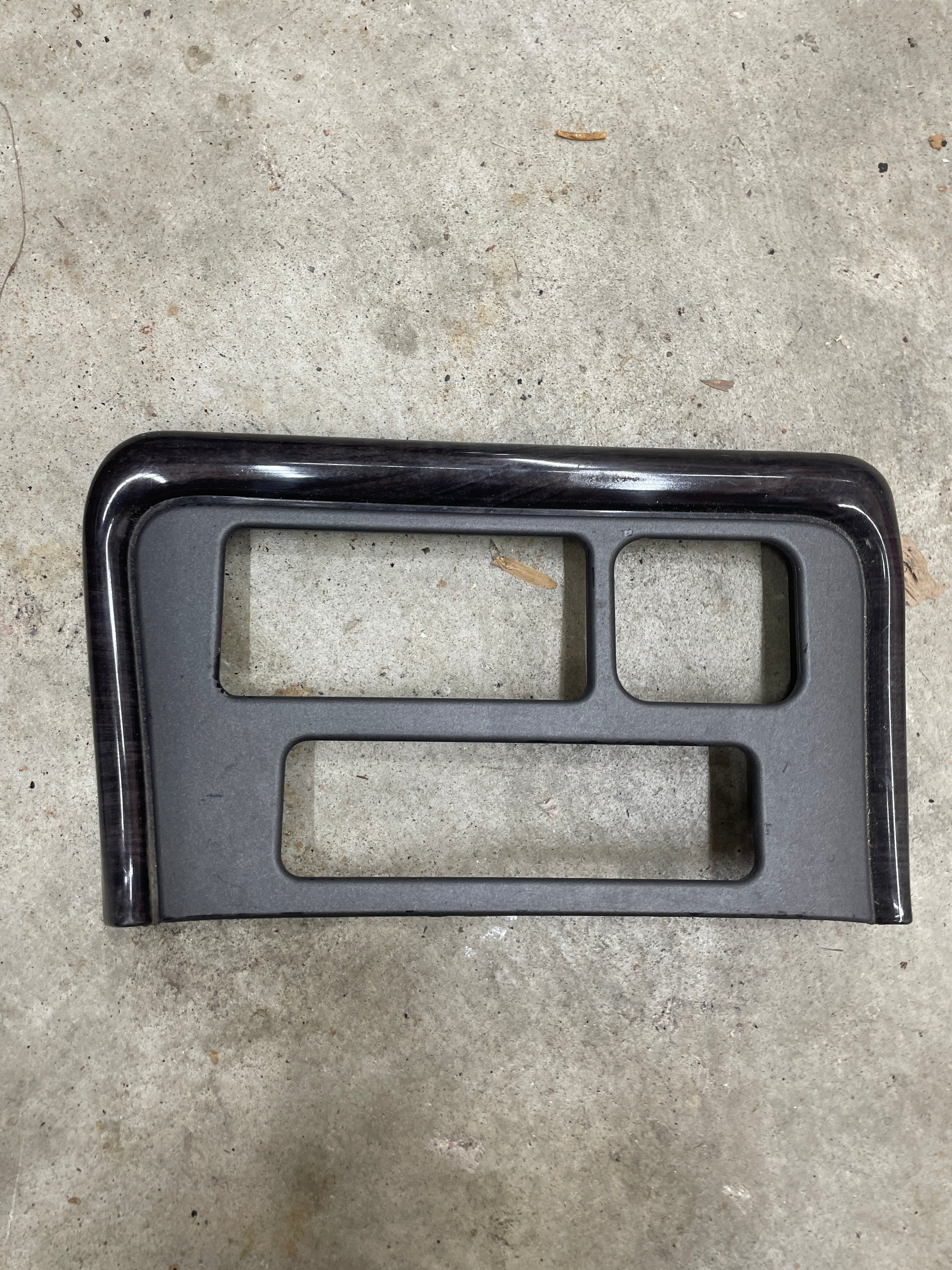 A General Motors OEM Center Console Front Bin Dark Woodgrain Trim for 2001-2002 GMC Sierra C3 or Yukon Denali is shown lying on a concrete floor.