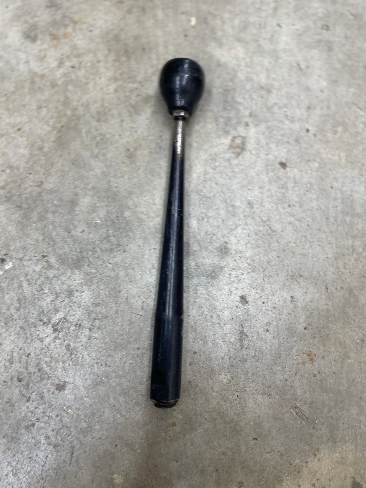 A General Motors OEM 5 Speed Manual Transmission Shifter Handle for 1982-1993 Chevy S10, Blazer, and more, with a black vintage look and round knob, lies on a gray concrete surface. The handle shows wear and tapers from the knob.