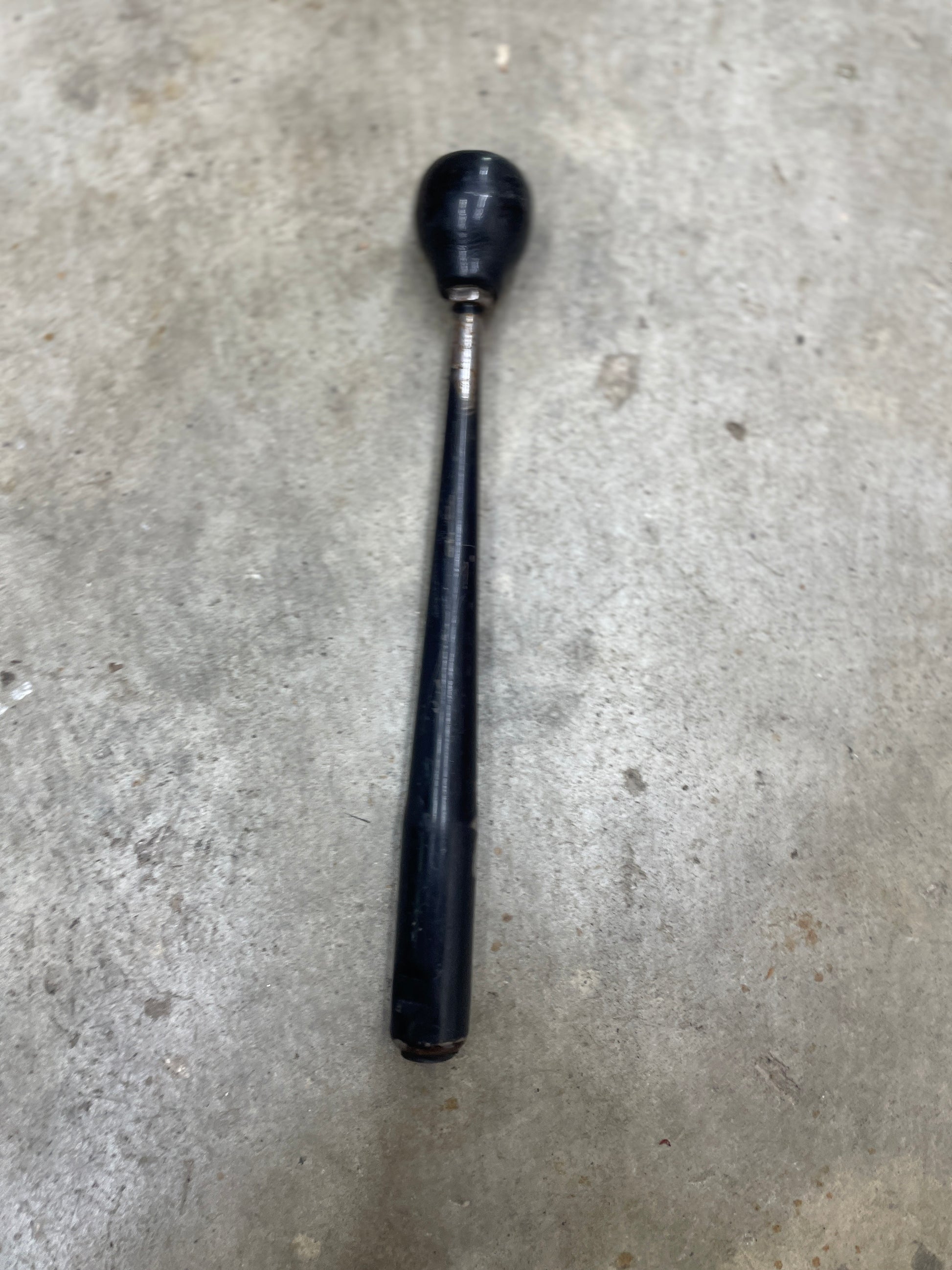 A General Motors OEM 5 Speed Manual Transmission Shifter Handle for 1982-1993 Chevy S10, Blazer, and more, with a black vintage look and round knob, lies on a gray concrete surface. The handle shows wear and tapers from the knob.