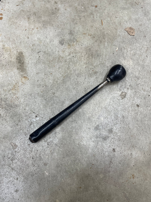 A General Motors OEM 5 Speed Manual Transmission Shifter Handle for 1982–1993 Chevy S10 and Blazer, black with a tapered, rounded end and shiny finish, is shown lying on rough concrete.