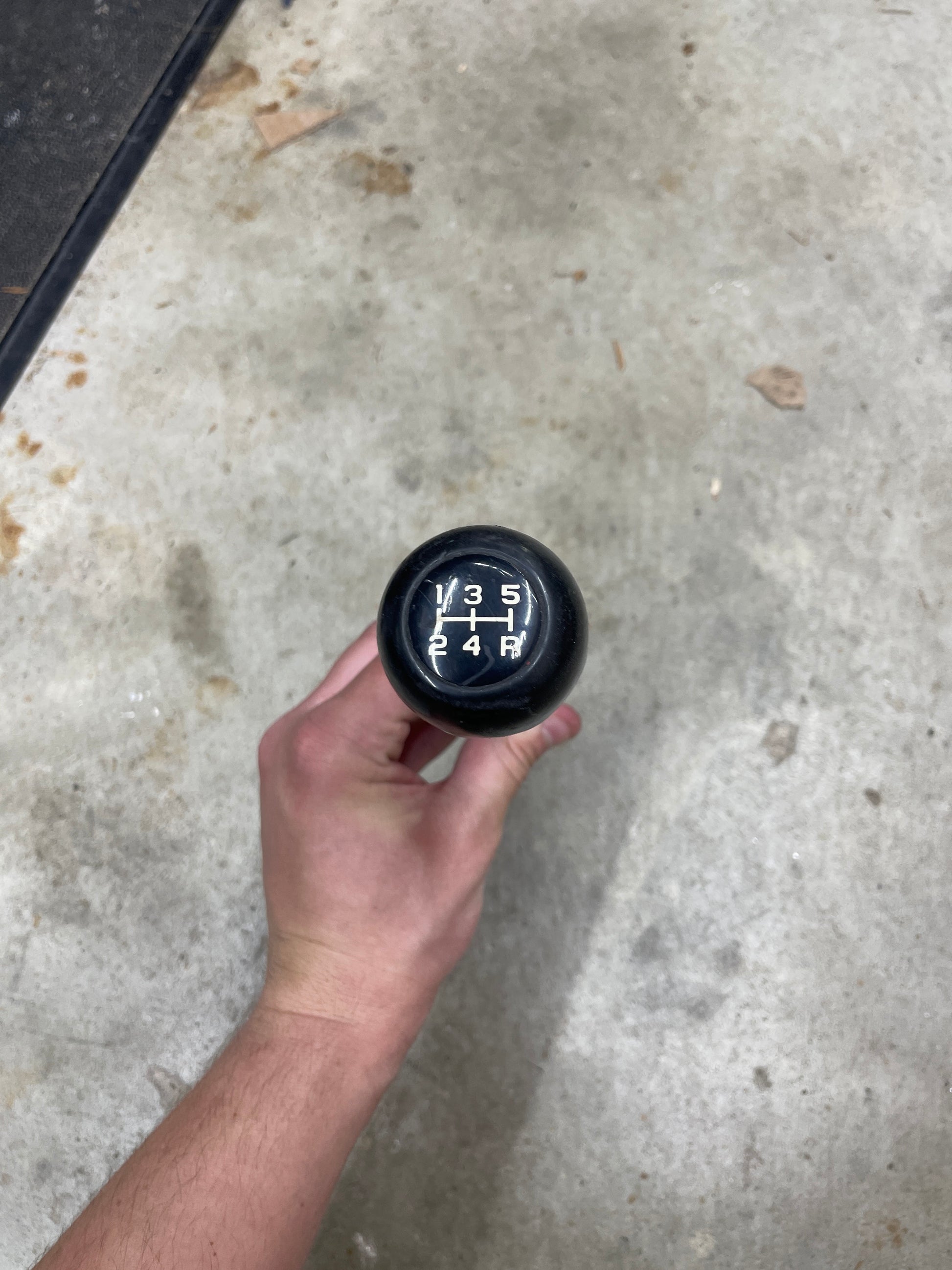 A hand grips a General Motors OEM 5 Speed Manual Transmission Shifter Handle for 1982-1993 Chevy S10, Blazer, and more, showing gears 1-5 and reverse against a concrete floor background.