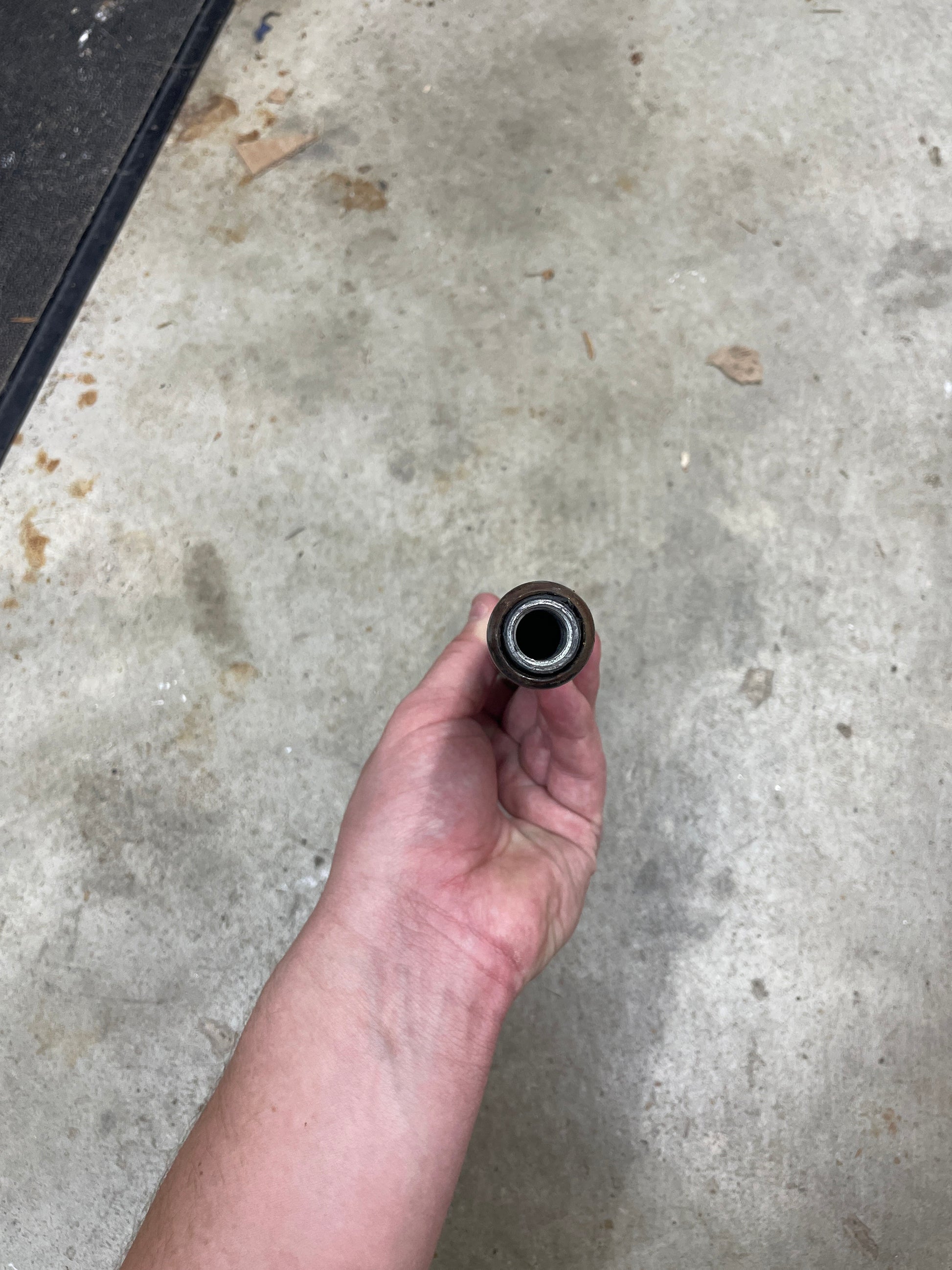 A hand holds a General Motors OEM 5 Speed Manual Transmission Shifter Handle for 1982–1993 Chevy S10, Blazer, and more, viewed from the end above a concrete floor with debris scattered around.