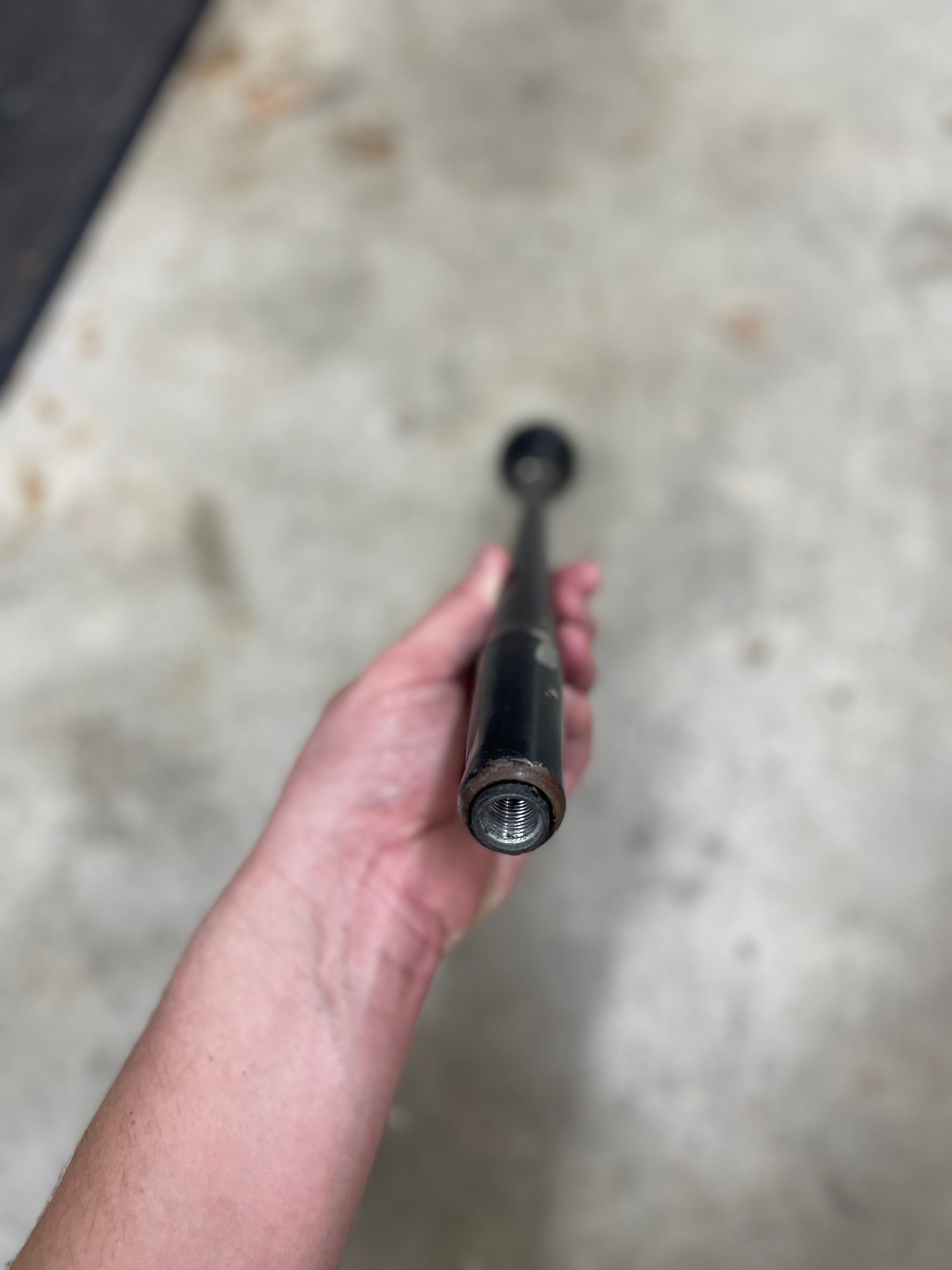 A hand holds a General Motors OEM 5 Speed Manual Transmission Shifter Handle for 1982-1993 Chevy S10, Blazer, and more, viewed from the front against a blurry light-colored concrete background.